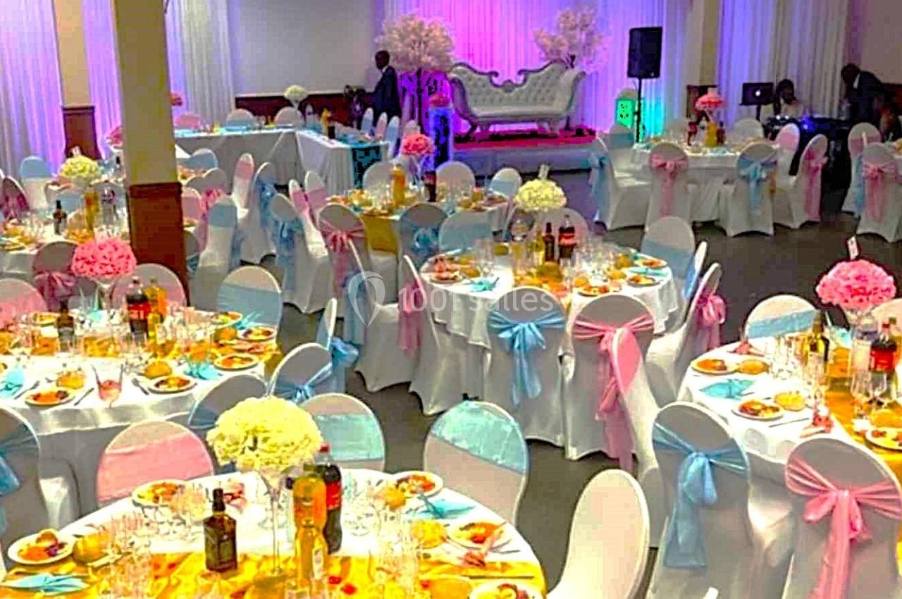 Salle de réception décorée avec des tables rondes ornées de nappes colorées, rubans et centres de table fleuris.