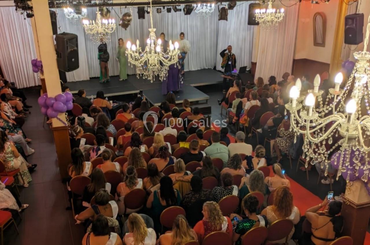 Salle comble avec un public assis face à une scène éclairée, décorée de lustres et de rideaux blancs.