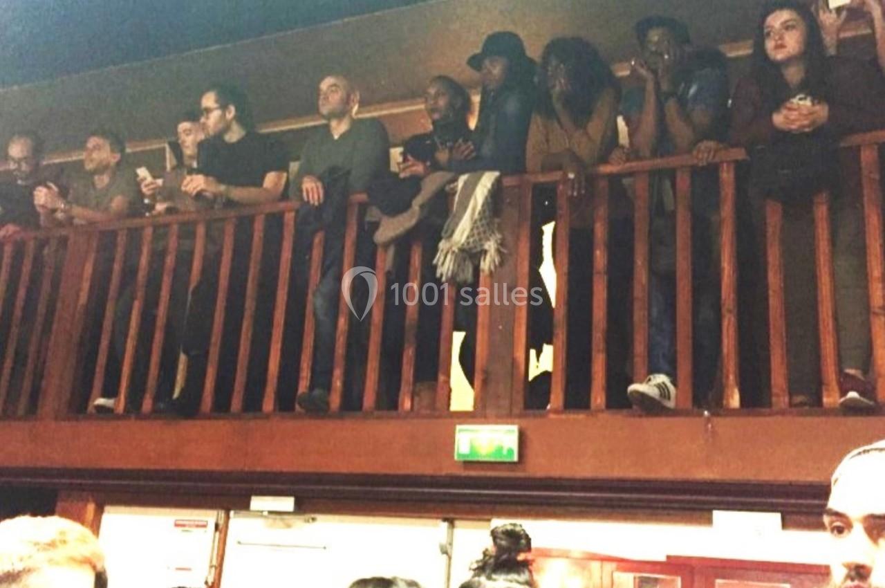 Des personnes debout sur une mezzanine en bois, observant une scène en contrebas dans un espace intérieur.