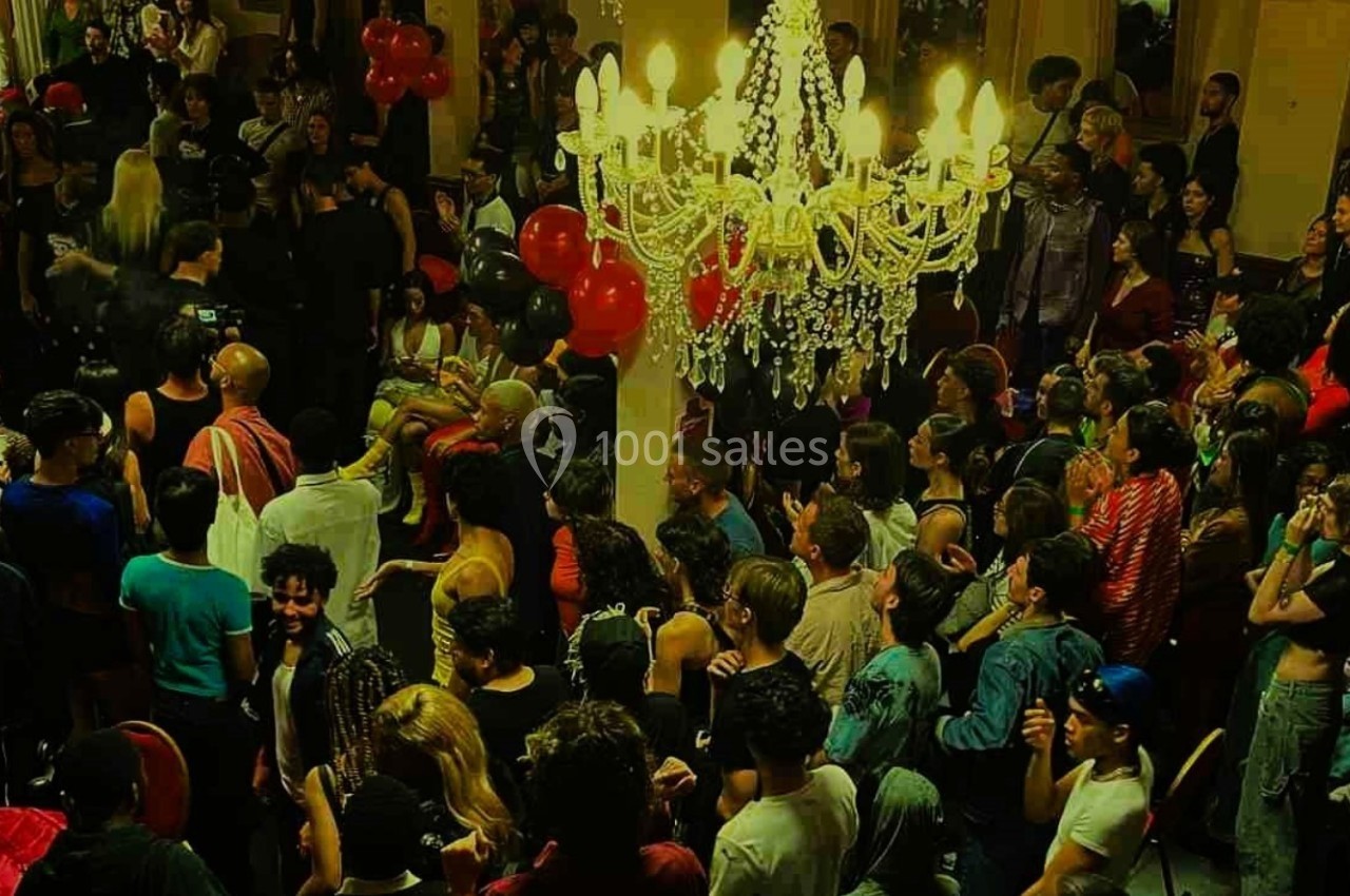 Foule rassemblée dans une salle éclairée par des lustres, avec des ballons rouges en décoration.