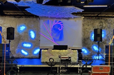 Salle sombre avec un public debout face à une scène éclairée où des musiciens jouent sous des projecteurs colorés.