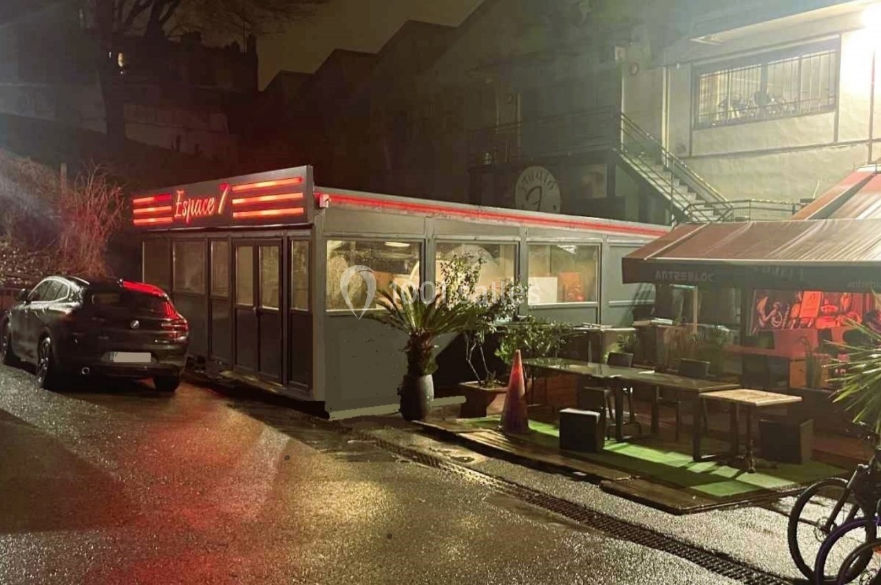 Extérieur d'un restaurant avec terrasse éclairée, tables en bois et plantes, dans une rue humide de nuit.