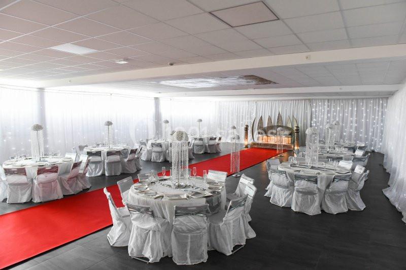 Salle de réception décorée avec des tables rondes dressées, nappes blanches, chandeliers et tapis rouge central.