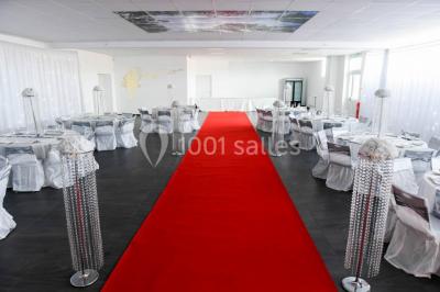 Salle de réception décorée avec des tables rondes dressées, nappes blanches, chandeliers et tapis rouge central.