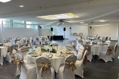Salle de réception décorée avec des tables rondes dressées, nappes blanches, chandeliers et tapis rouge central.