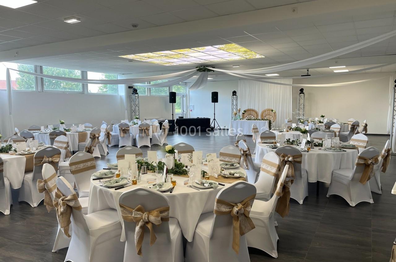 Salle de réception décorée pour un événement, avec tables rondes dressées et chaises ornées de nœuds en toile de jute.