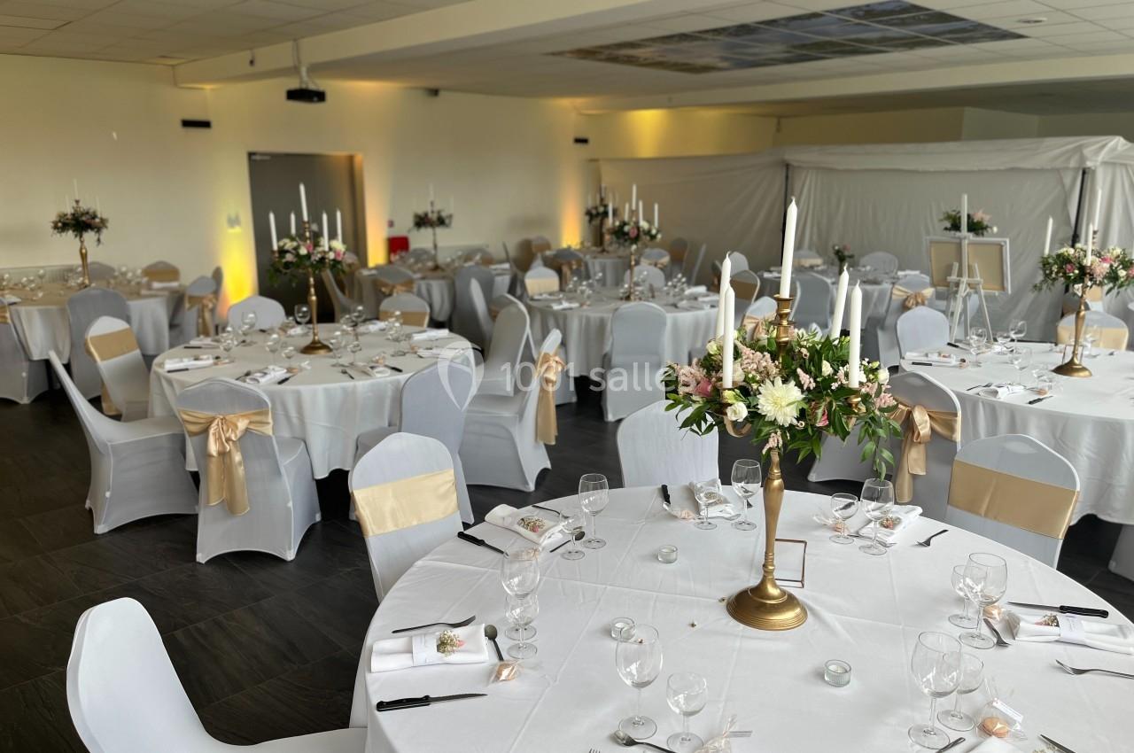 Salle de réception décorée avec des tables rondes, nappes blanches, chaises ornées de rubans dorés et centres de table…