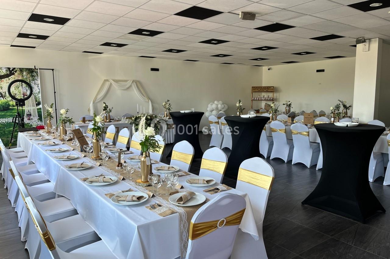 Salle de réception décorée avec des tables dressées, des chaises blanches ornées de rubans dorés et des éléments floraux.