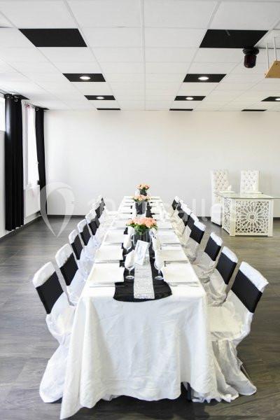Salle lumineuse avec une grande table rectangulaire dressée pour un repas, décorée de nappes blanches et de fleurs.