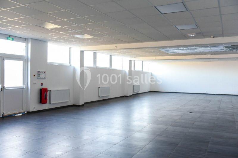 Salle vide avec sol en carrelage gris, murs blancs, grandes fenêtres laissant entrer la lumière naturelle.