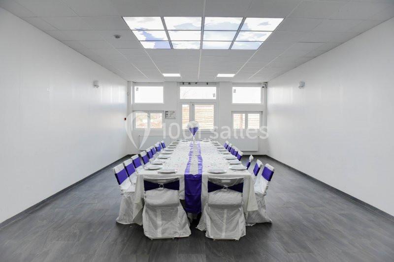 Salle lumineuse avec une grande table rectangulaire dressée pour un repas, chaises décorées de housses blanches et rubans…