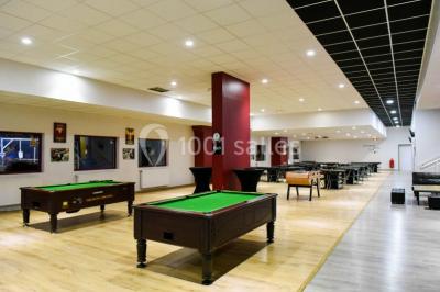 Salle de loisirs avec tables de billard, baby-foot, canapés noirs et murs rouges dans un espace lumineux.