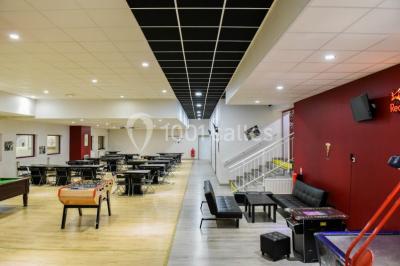 Salle de loisirs avec tables de billard, baby-foot, canapés noirs et murs rouges dans un espace lumineux.