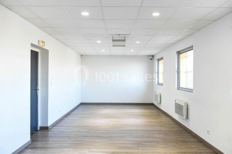 Salle vide avec parquet en bois, murs blancs, plafond à dalles et éclairage encastré, deux fenêtres et une porte.