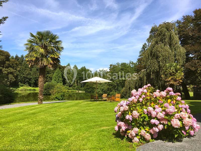 Jardin verdoyant avec pelouse, palmiers, hortensias en fleurs, table et chaises sous un parasol près d'un étang.