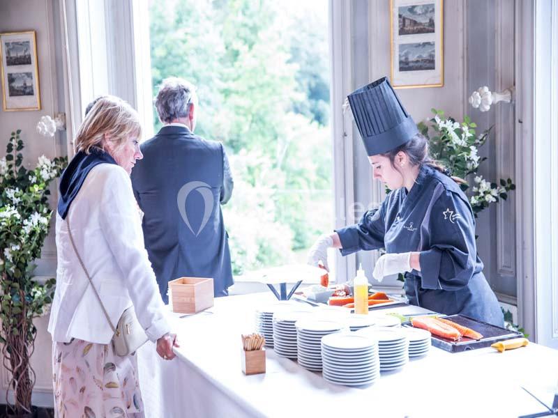 Une cheffe prépare des plats sur un buffet, observée par une femme, dans une salle lumineuse avec vue sur un jardin.