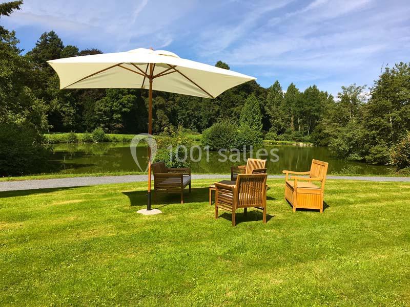 Mobilier de jardin en bois avec parasol blanc sur une pelouse, près d'un étang entouré d'arbres.