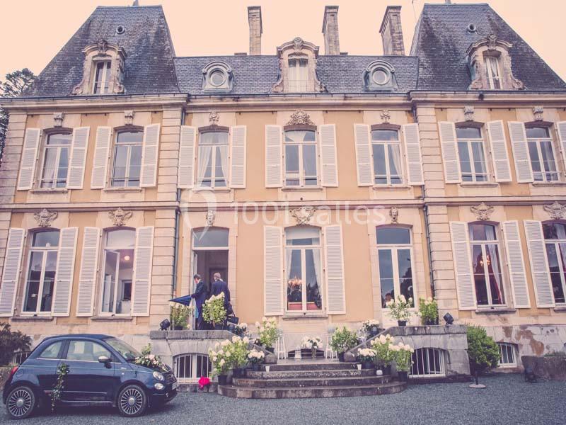 Façade d'un manoir ancien avec volets blancs, une petite voiture noire stationnée devant et des fleurs autour.