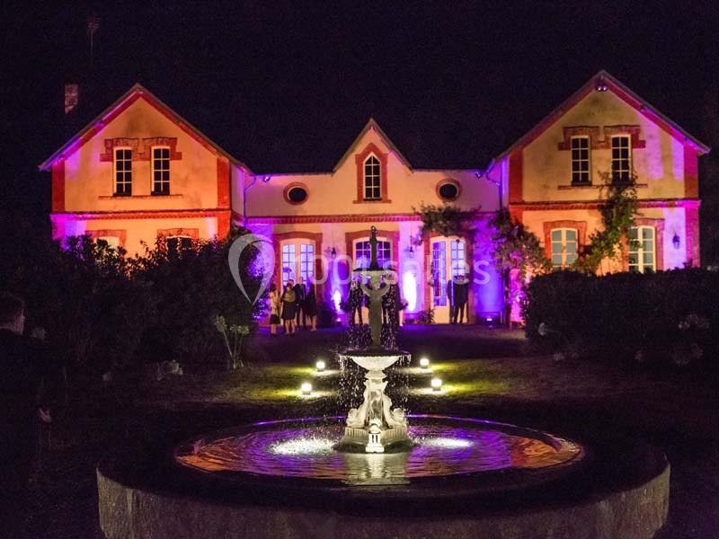 Façade d'une maison éclairée par des lumières colorées avec une fontaine illuminée au premier plan, de nuit.