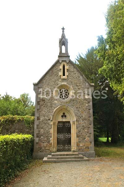 Petite chapelle en pierre avec une porte ornée et un vitrail, entourée de verdure et éclairée par une lumière naturelle.