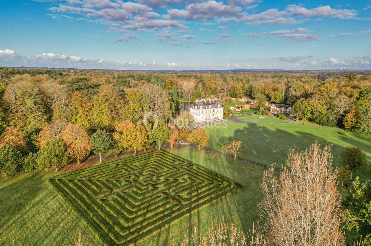 Vue aérienne d'un château entouré de jardins, d'un labyrinthe végétal et d'une forêt en arrière-plan.