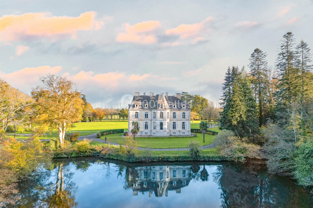Château entouré d'arbres, avec un étang reflétant sa façade sous un ciel partiellement nuageux.