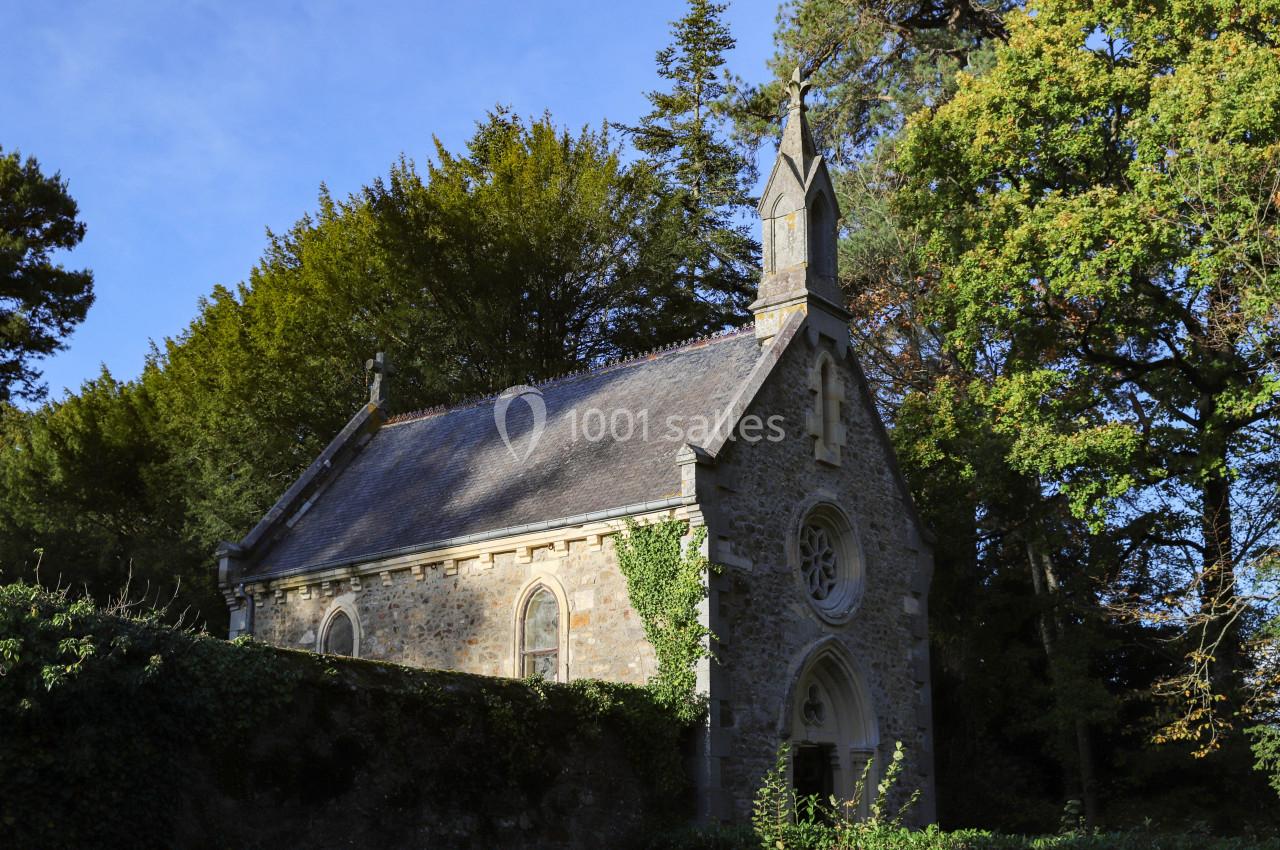 Petite chapelle en pierre entourée d'arbres, avec un toit en ardoise et une façade ornée d'une rosace.