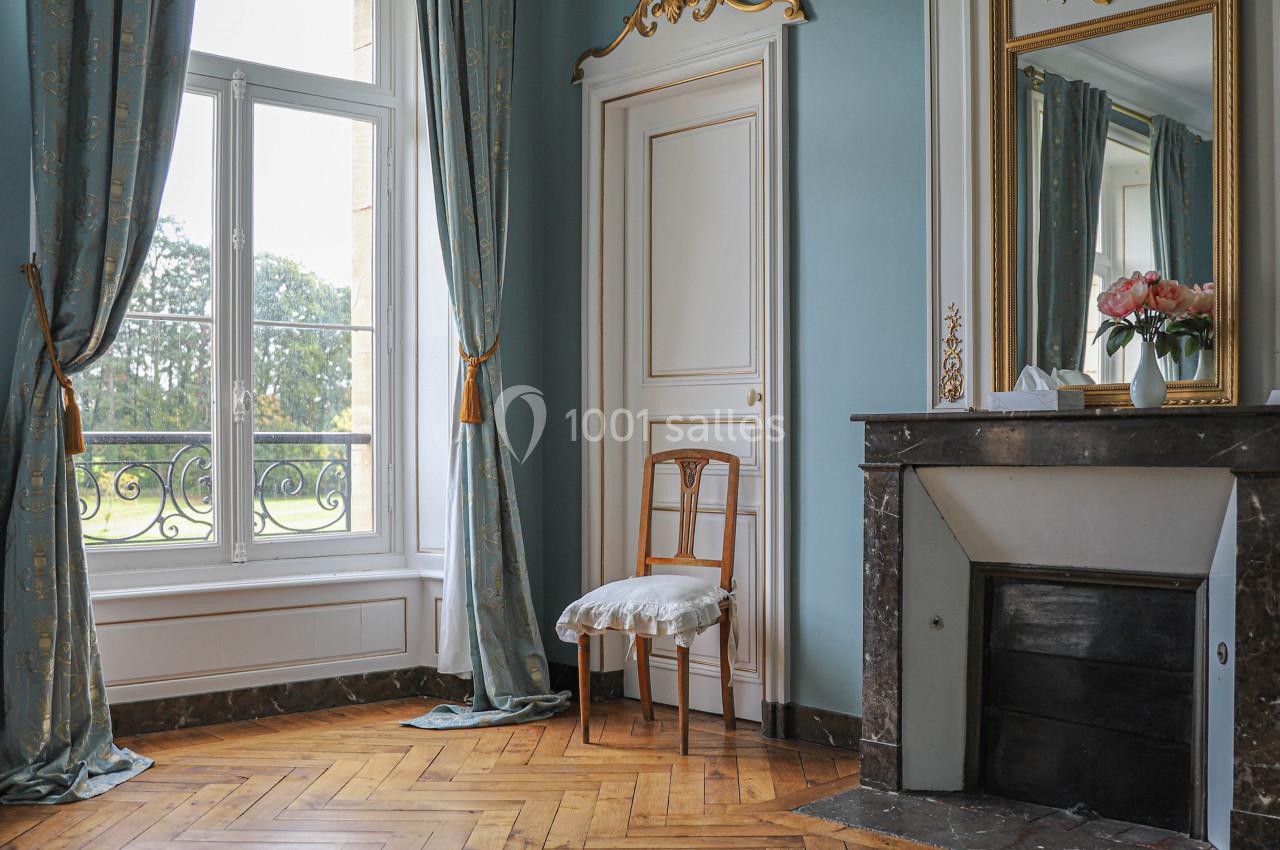 Pièce lumineuse avec parquet en bois, cheminée en marbre, miroir doré, rideaux bleus et vue sur un jardin.