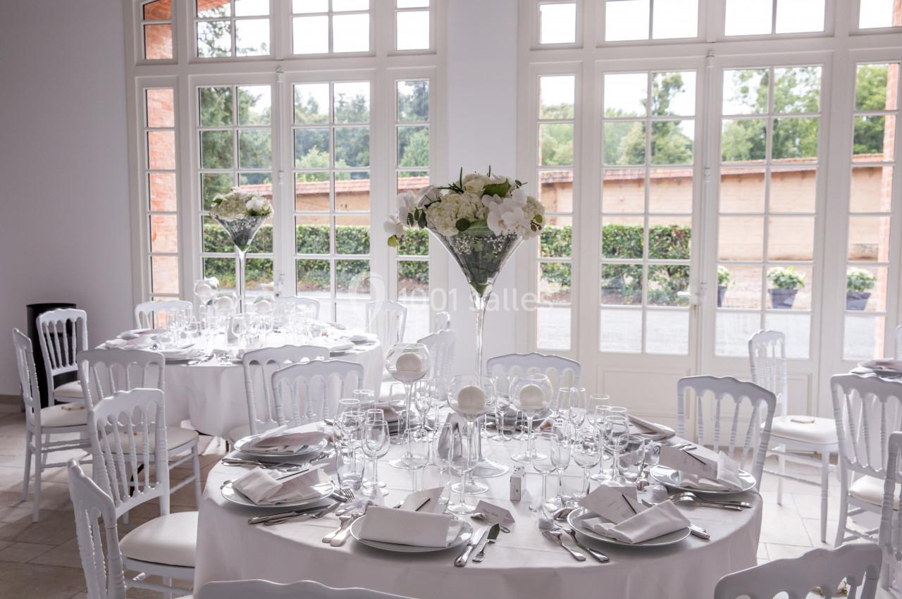 Salle lumineuse avec tables rondes dressées élégamment, décorées de fleurs blanches et entourées de chaises blanches.