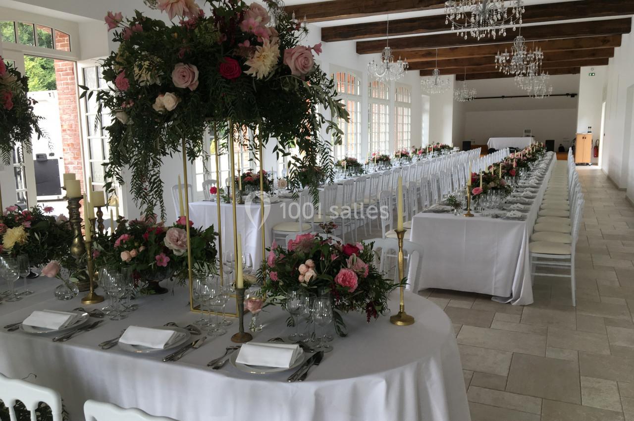 Salle de réception lumineuse avec tables décorées de fleurs colorées, nappes blanches et chandeliers dorés.