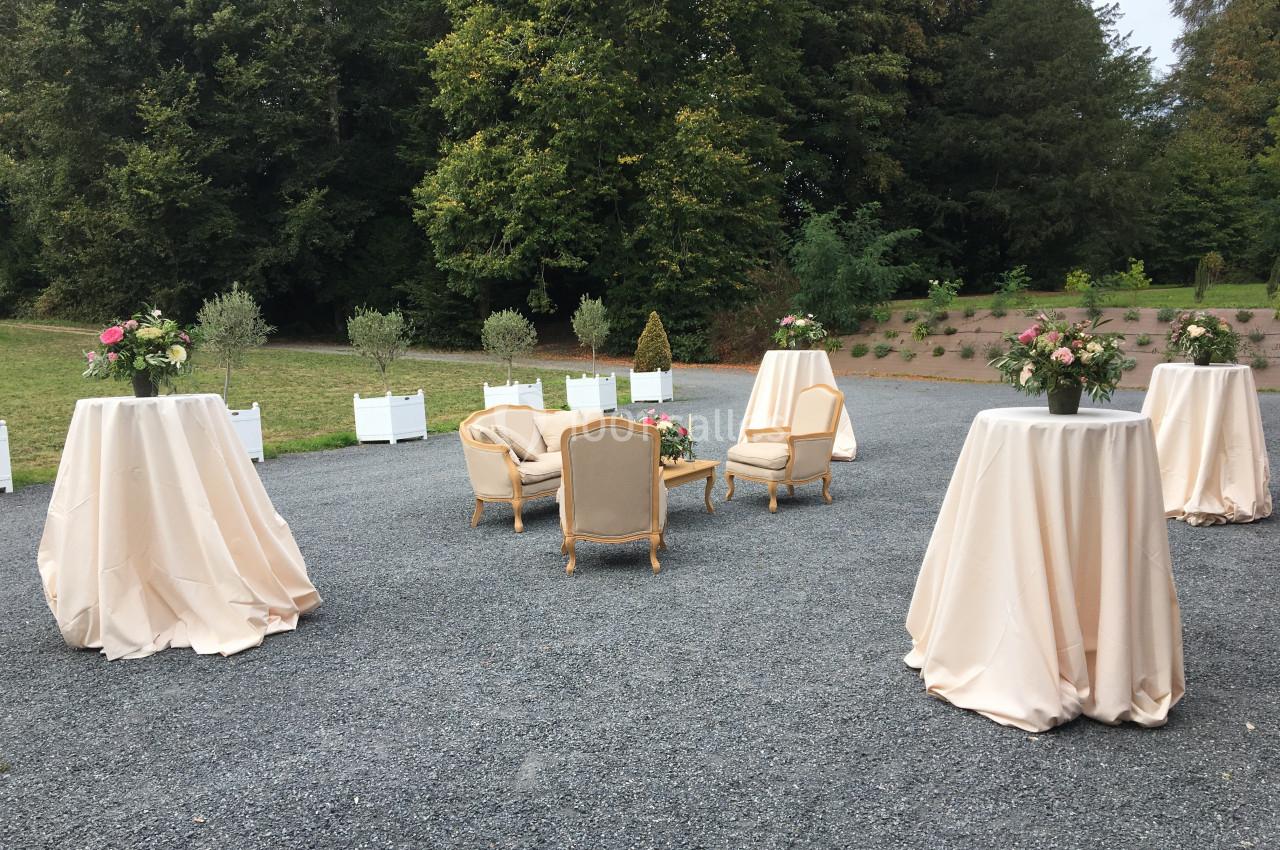 Mobilier de réception avec fauteuils et tables hautes drapées, disposés en extérieur sur un sol gravillonné.