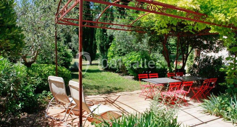 Location salle Fourques (Gard) - Domaine de la Tourette #11 Terrasse ombragée avec pergola rouge, chaises longues et table entourée de chaises rouges dans un jardin verdoyant.