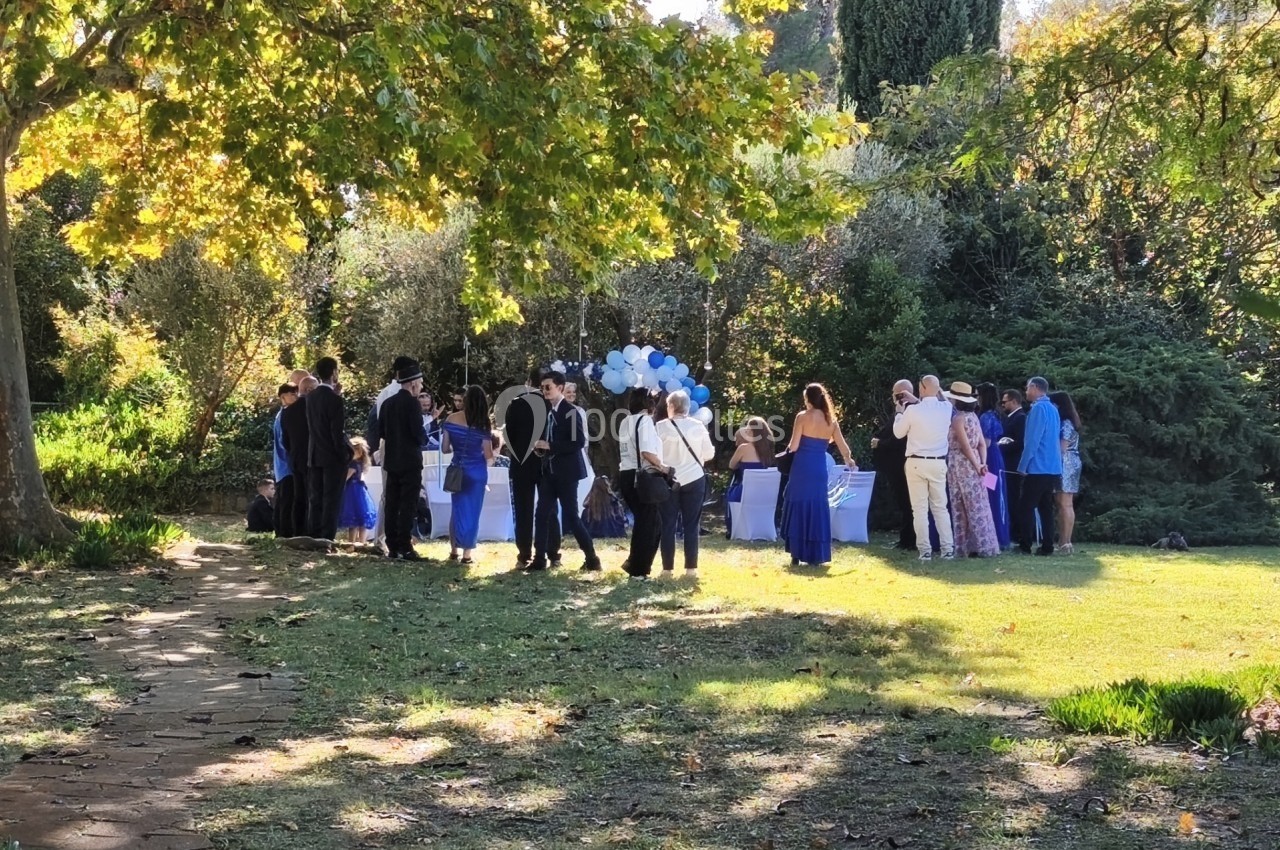 Groupe de personnes rassemblées dans un jardin ensoleillé, entourées d'arbres et de ballons bleus et blancs.