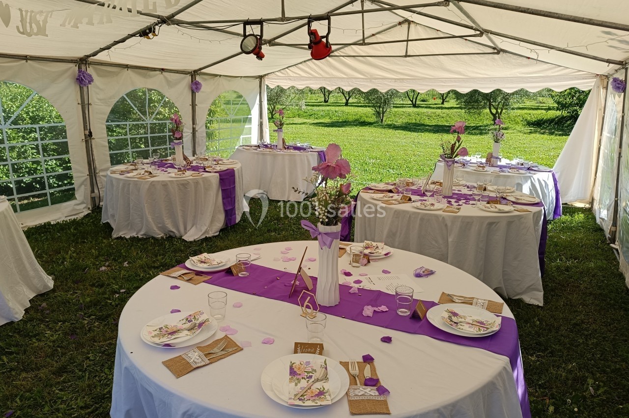 Tables rondes décorées en blanc et violet sous une tente, disposées pour un événement en plein air.