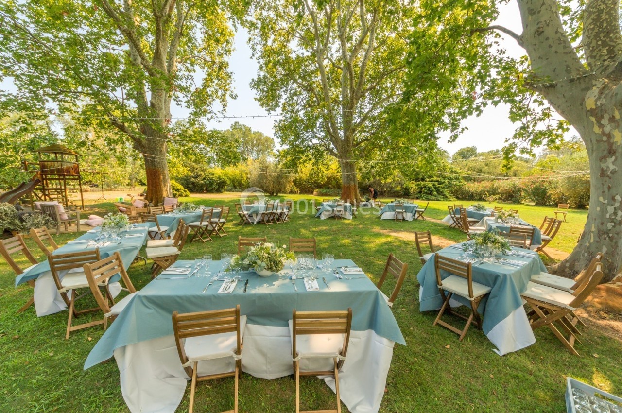 Tables dressées avec nappes bleues et blanches, disposées en extérieur sous de grands arbres dans un cadre verdoyant.