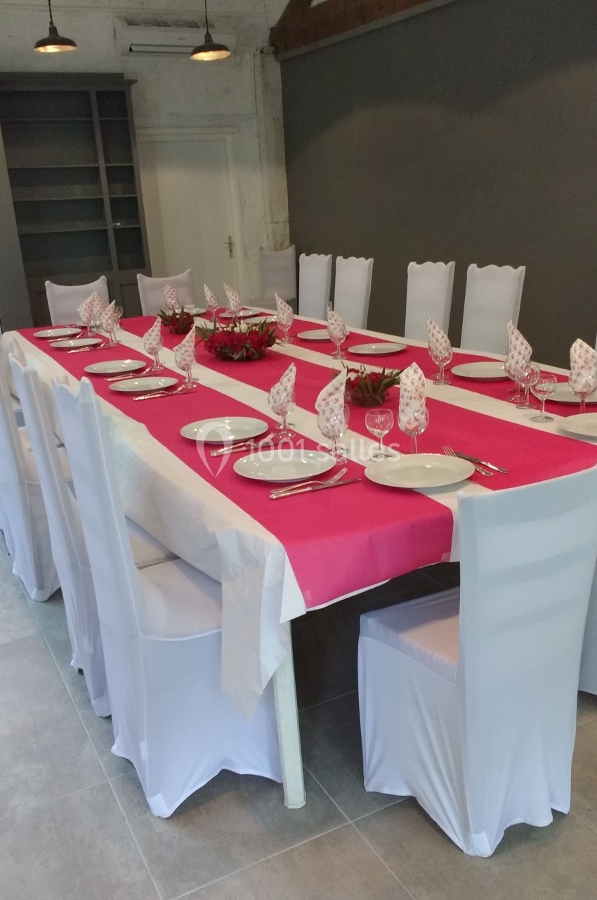 Table dressée avec une nappe rose et blanche, assiettes, verres et serviettes pliées, entourée de chaises blanches.