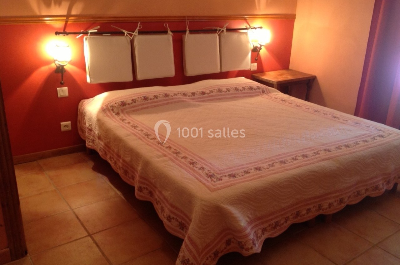 Lit double avec couvre-lit rose et blanc dans une chambre aux murs rouges et orange, éclairée par deux appliques murales.