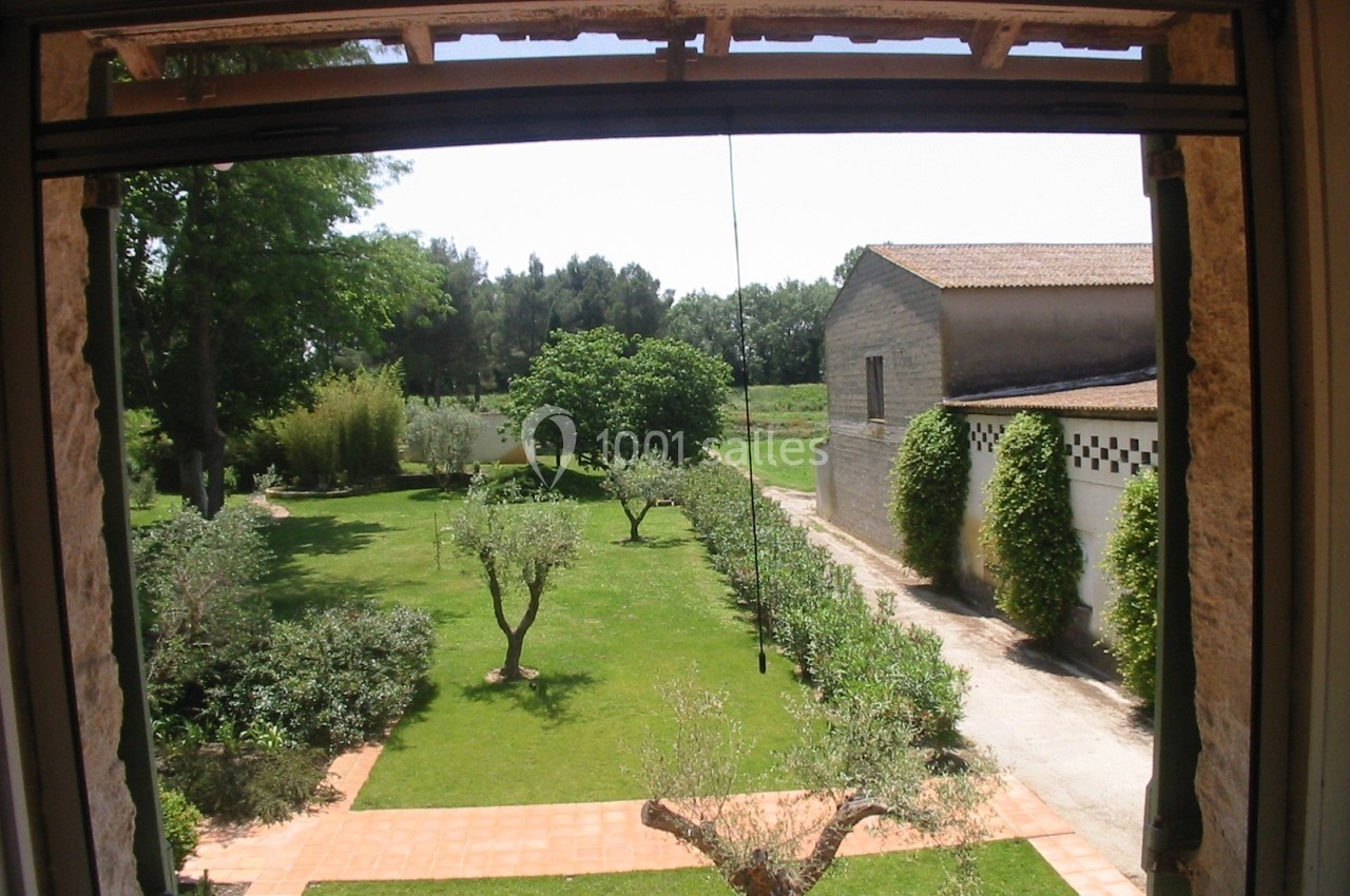 Vue depuis une fenêtre sur un jardin arboré avec des allées pavées, des oliviers et un bâtiment en pierre.
