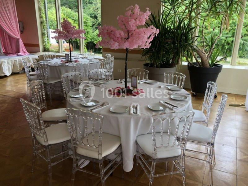 Salle décorée pour un événement avec tables rondes, nappes blanches, chaises transparentes et centres de table fleuris.
