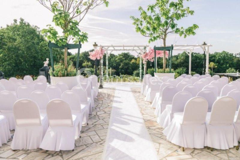 Allée centrale bordée de chaises blanches menant à une arche décorée de fleurs roses dans un cadre extérieur verdoyant.
