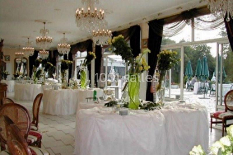 Salle de réception lumineuse avec tables décorées de nappes blanches, chandeliers et arrangements floraux verts.