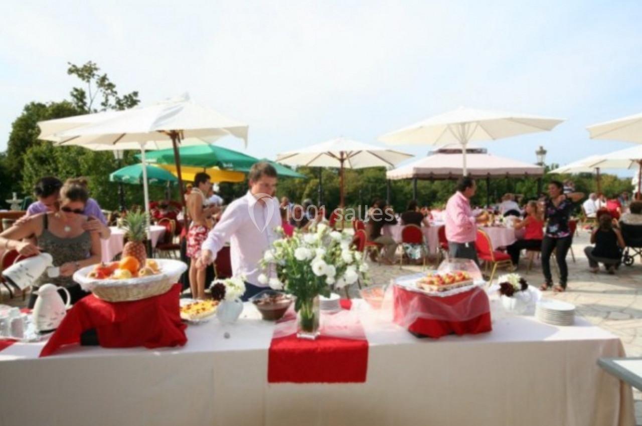 Buffet en plein air avec nappes blanches et rouges, fleurs au centre, invités se servant sous des parasols.
