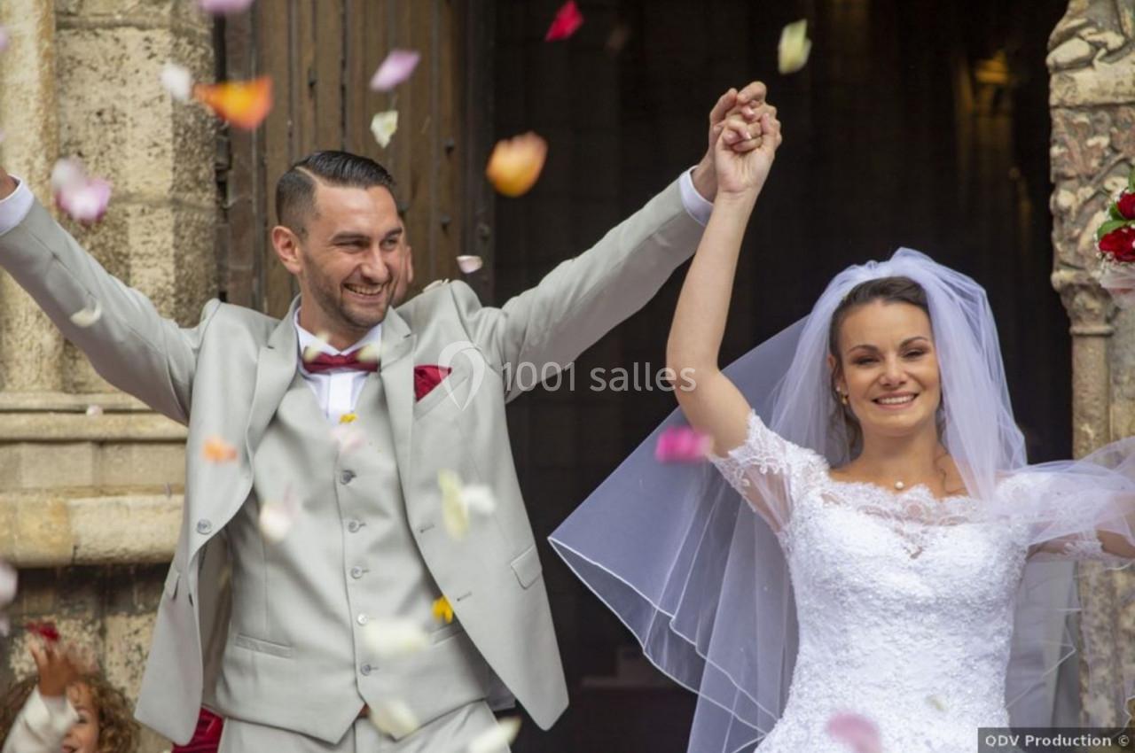 Un couple de mariés souriant sort d'une église sous une pluie de pétales colorés.