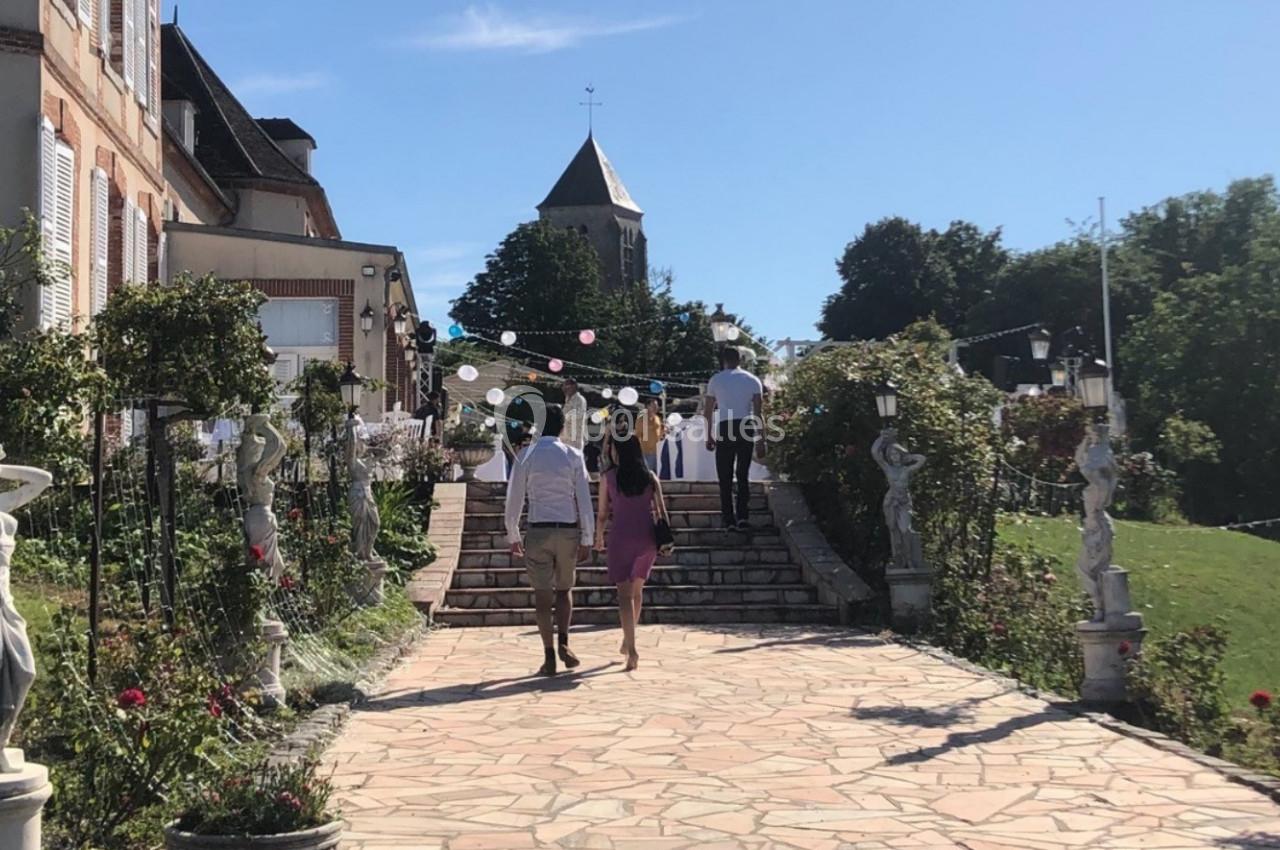 Allée pavée bordée de statues et de fleurs menant à un bâtiment ancien avec une église en arrière-plan.