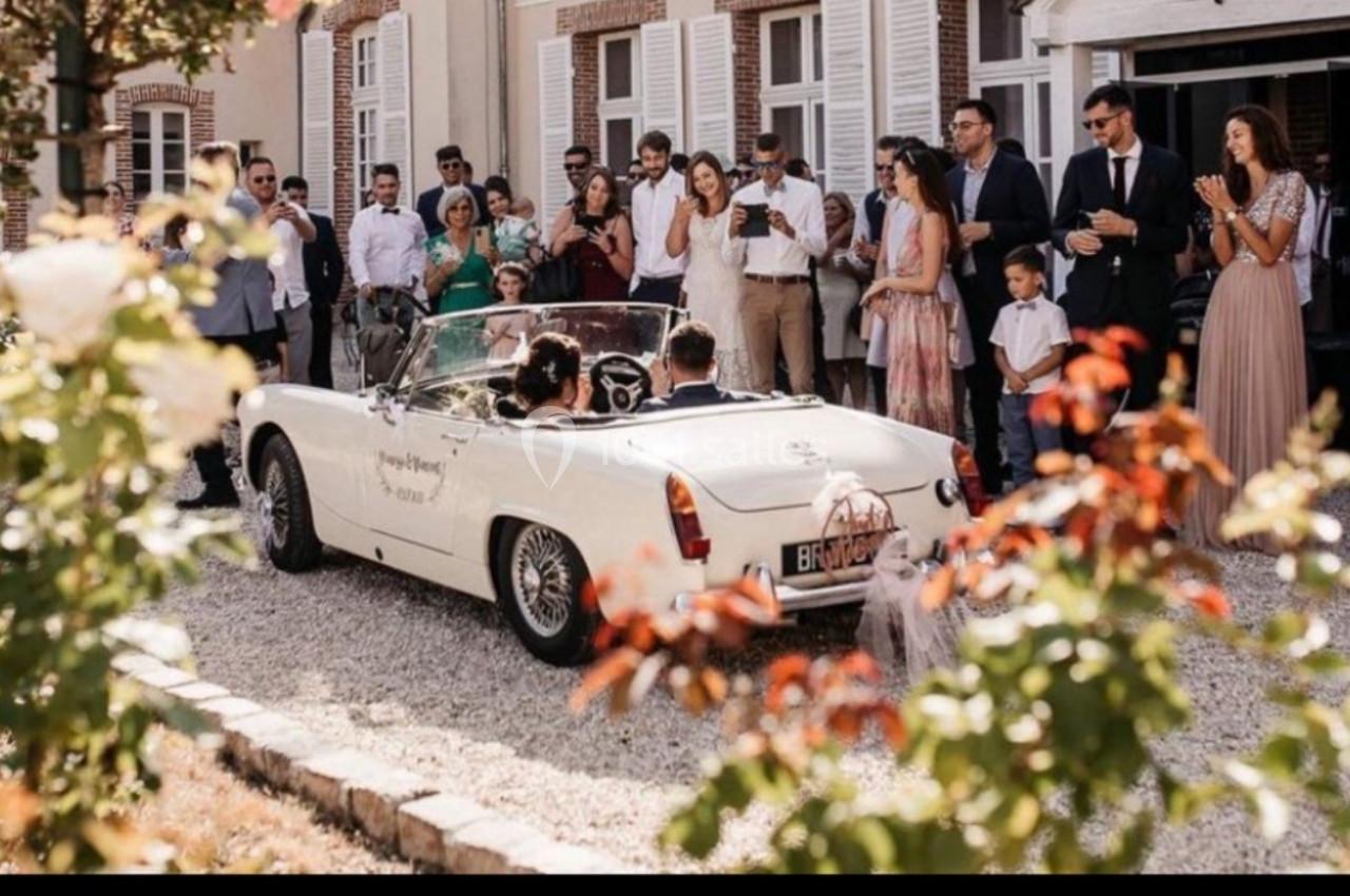Un couple dans une voiture décapotable blanche quitte une cérémonie entouré d'invités élégants devant un bâtiment.