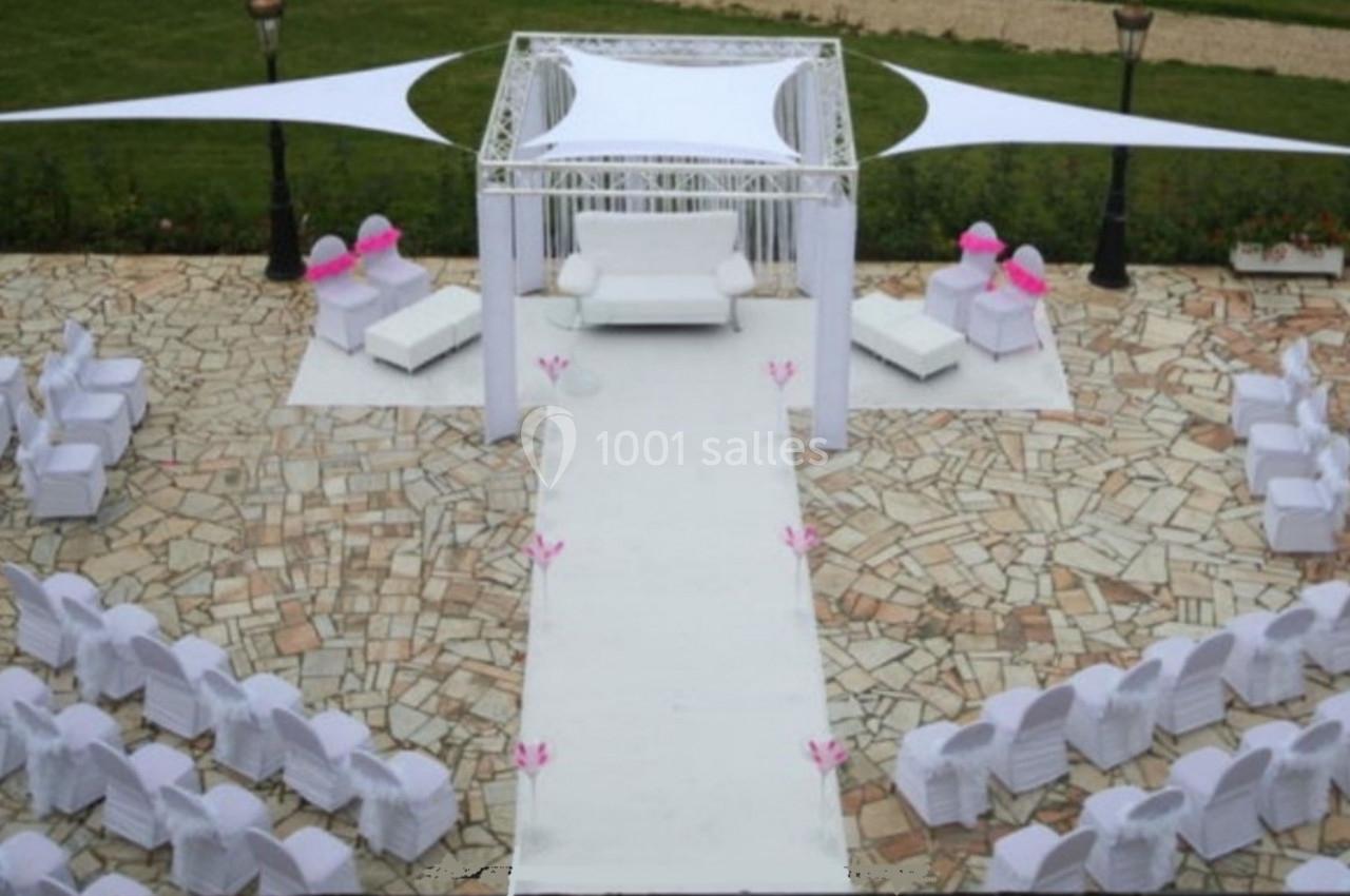 Allée centrale décorée pour une cérémonie en extérieur, entourée de chaises blanches et d'un espace couvert.