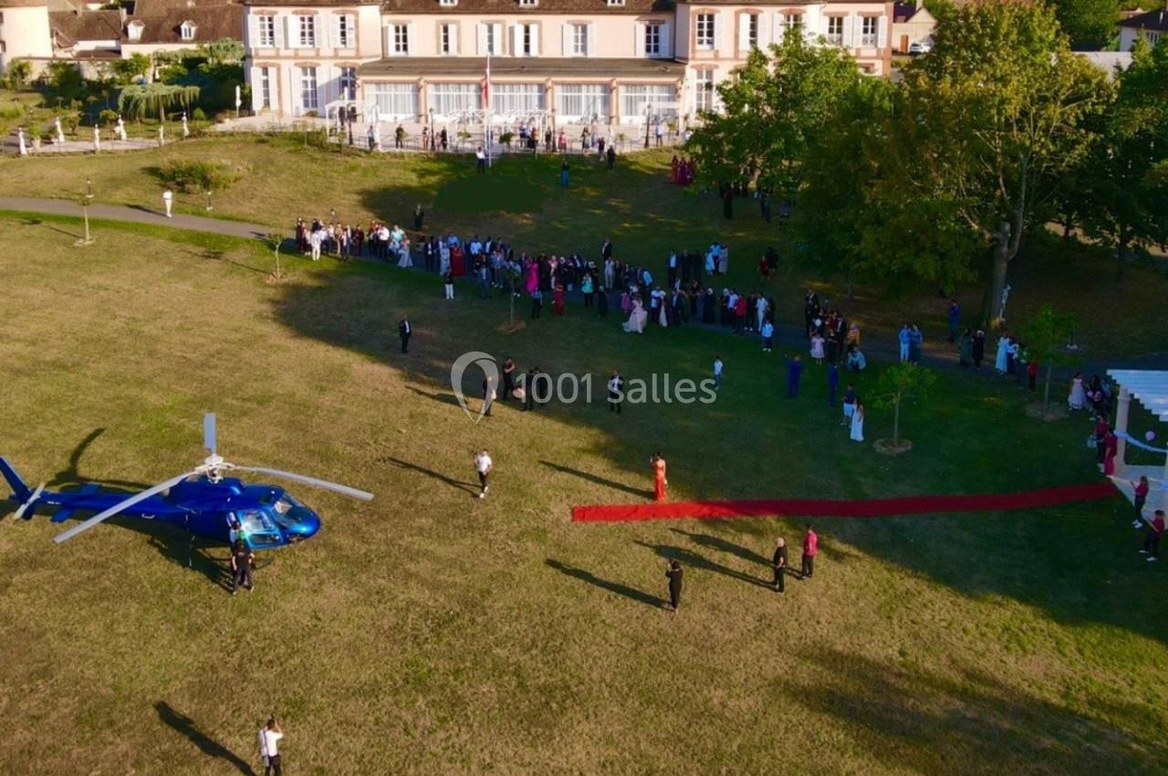 Vue aérienne d'un hélicoptère bleu posé sur une pelouse près d'un bâtiment, avec des invités alignés sur un tapis rouge.