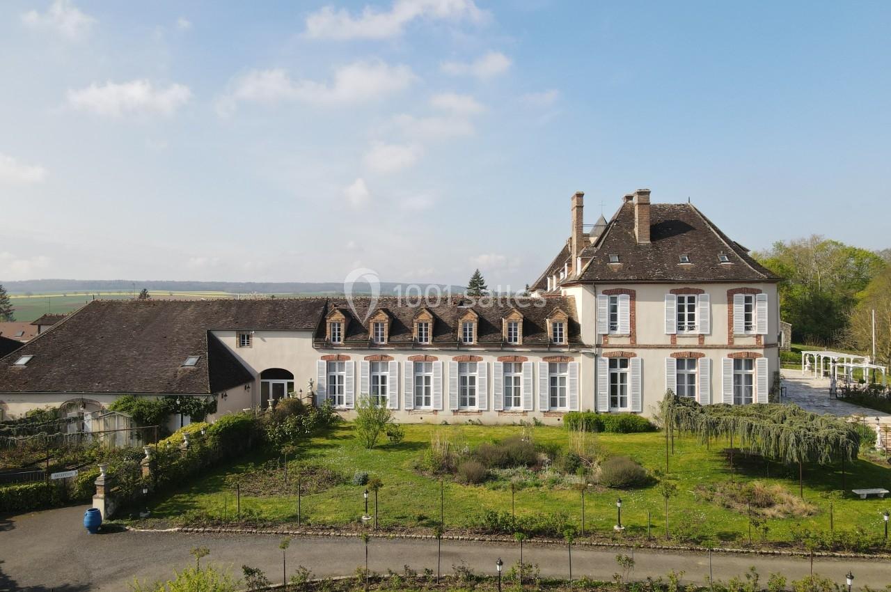 Vue d'un grand bâtiment ancien avec des fenêtres en arc, entouré de jardins et d'un paysage rural sous un ciel dégagé.