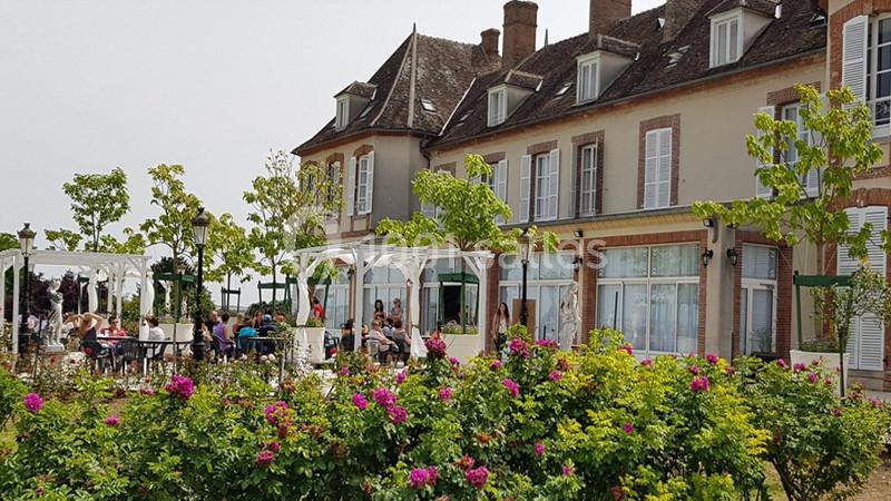Façade d'un bâtiment ancien avec terrasse animée, entourée de rosiers et d'arbres sous un ciel clair.