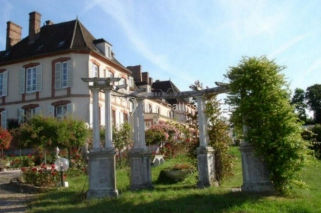 Jardin verdoyant avec pergolas en pierre devant une grande maison de style traditionnel sous un ciel dégagé.